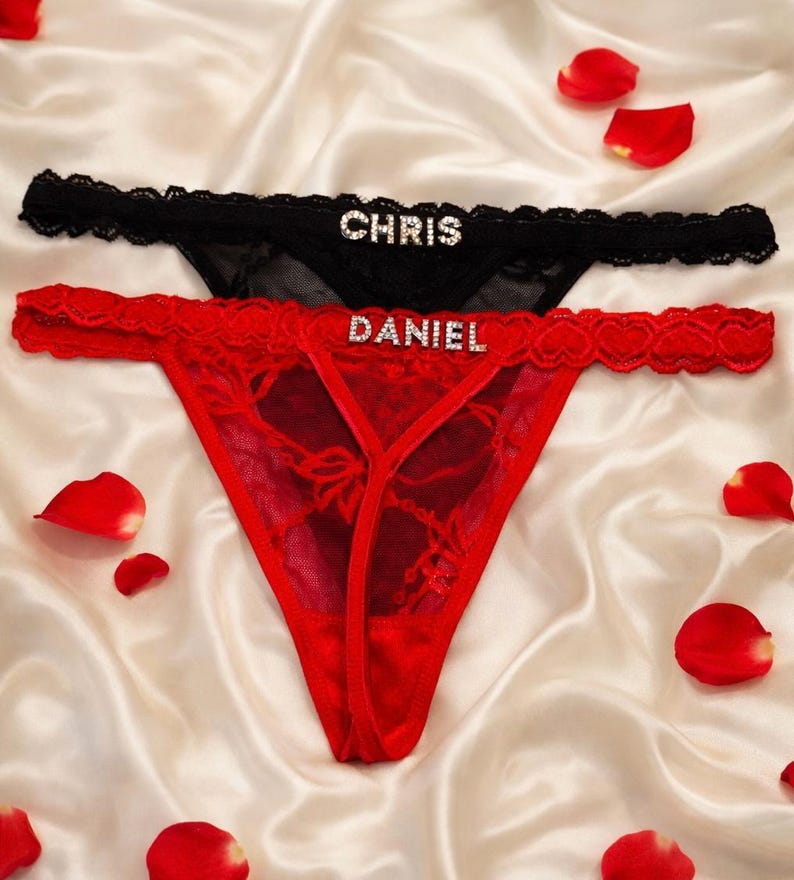 Peut inclure: Deux strings, un noir et un rouge, avec des d&eacute;tails en dentelle. Le string noir porte le nom "CHRIS" en strass, et le string rouge porte "DANIEL". Des p&eacute;tales de roses rouges sont dispers&eacute;s autour des sous-v&ecirc;tements.