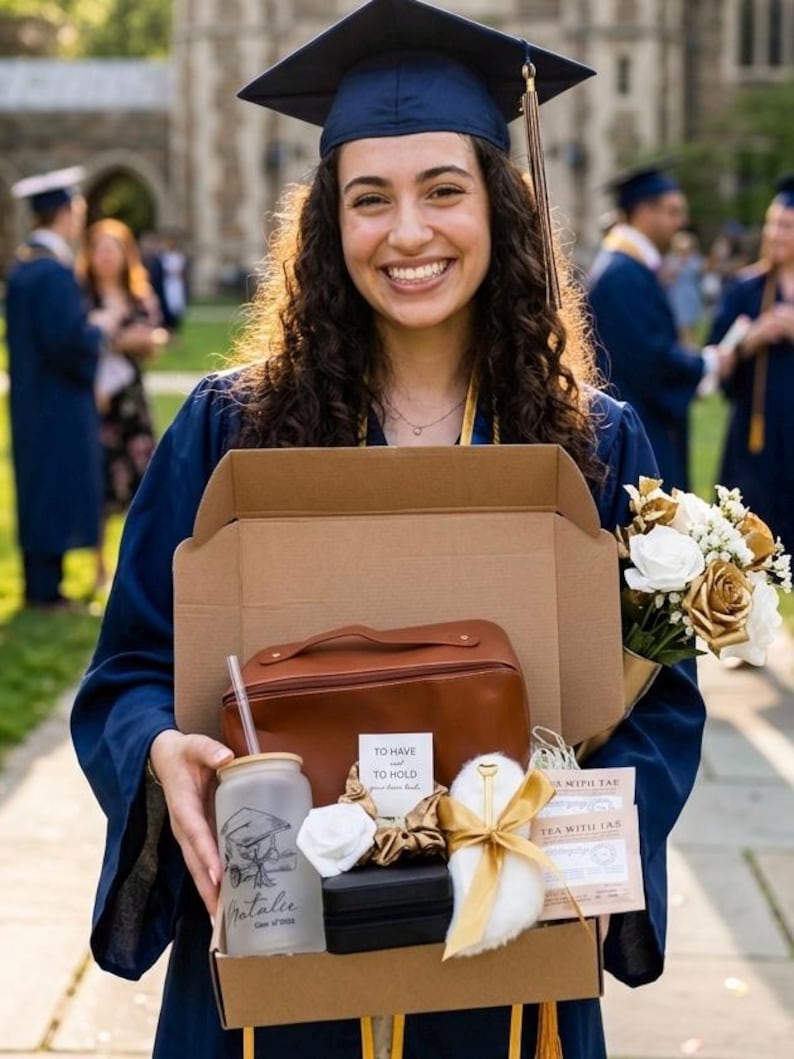 May include: A graduate in a navy blue cap and gown smiles, holding a gift box with a brown leather bag, a personalized glass, and a bouquet of white and gold flowers. The glass is inscribed with "Natalie" and "Class of 2024".