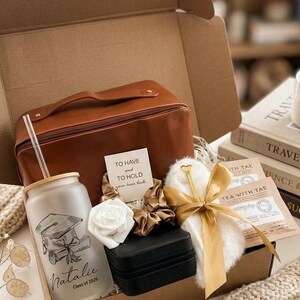 May include: A gift box containing a brown leather-look travel case, a frosted glass tumbler with a bamboo lid and straw featuring a graduation cap graphic and the name Natalie Class of 2026, a black jewellery box, a white faux fur item, and tea.