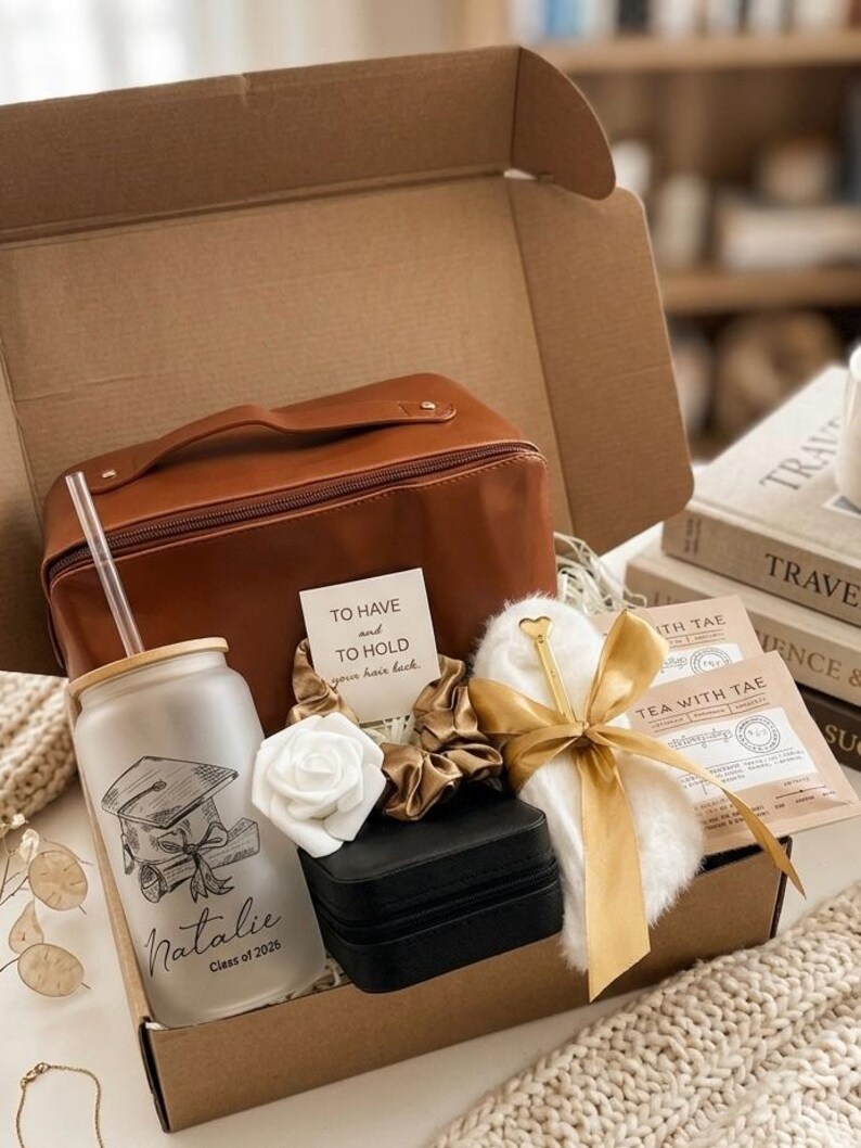 May include: A gift box containing a brown leather-look travel case, a frosted glass tumbler with a bamboo lid and straw featuring a graduation cap graphic and the name Natalie Class of 2026, a black jewellery box, a white faux fur item, and tea.