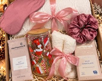 May include: A gift box containing a pink satin sleep mask, a white knit blanket tied with a pink ribbon, a pink satin scrunchie, a glass tumbler with a bamboo lid and holiday print, fuzzy white socks, and two Moonstruck milk chocolate bars.