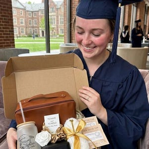 May include: A cardboard gift box containing a brown leather-effect toiletry bag, a frosted glass tumbler with a straw featuring a graduation cap graphic and the name Natalie, a white rose, gold scrunchies, a black jewellery case, and tea packets.