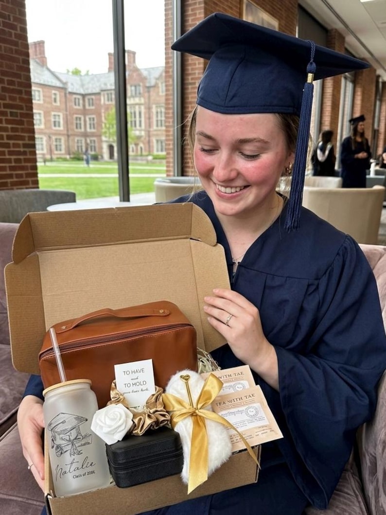 May include: A cardboard gift box containing a brown leather-effect toiletry bag, a frosted glass tumbler with a straw featuring a graduation cap graphic and the name Natalie, a white rose, gold scrunchies, a black jewellery case, and tea packets.