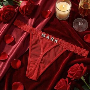 Peut inclure: String rouge avec le nom "MARK" en lettres argent&eacute;es. Le sous-v&ecirc;tement a une bordure en dentelle et est pr&eacute;sent&eacute; sur un tissu de velours rouge avec des p&eacute;tales et des roses.