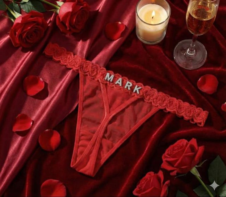 Peut inclure: String rouge avec le nom "MARK" en lettres argent&eacute;es. Le sous-v&ecirc;tement a une bordure en dentelle et est pr&eacute;sent&eacute; sur un tissu de velours rouge avec des p&eacute;tales et des roses.