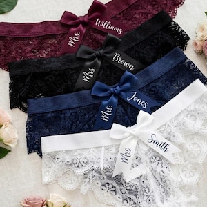 May include: A selection of lace underwear in shades of burgundy, black, navy, and white, each with a satin ribbon bow. The bows are personalised with names such as "Williams", "Brown", "Jones", and "Smith", and "Mrs."
