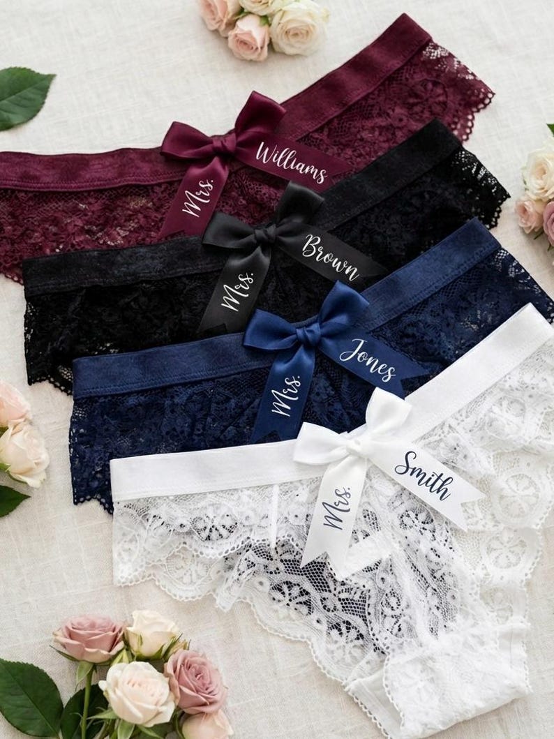 May include: A selection of lace underwear in shades of burgundy, black, navy, and white, each with a satin ribbon bow. The bows are personalised with names such as "Williams", "Brown", "Jones", and "Smith", and "Mrs."