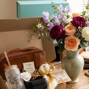 May include: A gift box containing a brown leather-look toiletry bag, a frosted glass tumbler with a graduation cap graphic and the name Natalie, a small black jewellery case, a white scrunchie with a gold ribbon, and a card reading To Have and To Hold.