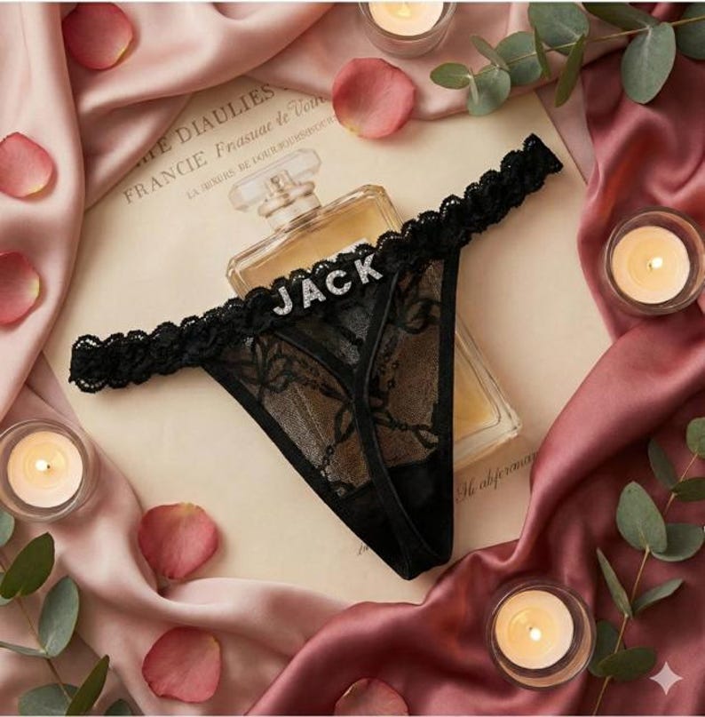 Peut inclure: String noir en dentelle avec le nom "JACK" en lettres argent&eacute;es. La lingerie a une bordure festonn&eacute;e et est pr&eacute;sent&eacute;e sur un livre vintage avec un flacon de parfum et des bougies allum&eacute;es en arri&egrave;re-plan.