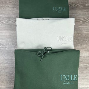 May include: Three green hooded sweatshirts stacked on top of each other. The top sweatshirt has the text "UNCLE Ben" embroidered on the front. The middle sweatshirt has the text "UNCLE Sean" embroidered on the front. The bottom sweatshirt has the text "UNCLE Andrew" embroidered on the front.