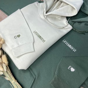 May include: Two hooded sweatshirts, one in a light beige color and the other in a dark green color. The beige sweatshirt has the Roman numeral LXVII.MMXVI embroidered on the front, and the letter C with a heart above it on the sleeve. The green sweatshirt has the Roman numeral LXVII.MMXVI embroidered on the front and the letter E with a heart above it on the sleeve.