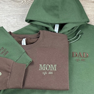 May include: Three sweatshirts with embroidered text. A brown sweatshirt with "MOM est. 2024" embroidered on the front. A green sweatshirt with "DAD est. 2024" embroidered on the front. A green sweatshirt with "KYLIE, HAILEY, SADIE" embroidered on the front.