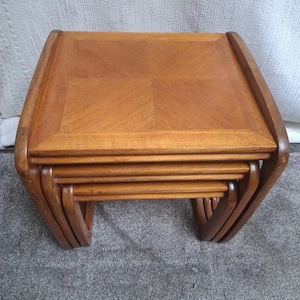 May include: A set of four wooden nesting tables in a light brown hue. Each table features a square top with a patterned surface and curved legs. The tables are designed to stack, with the smallest fitting inside the largest. The tables are approximately 50 cm in height.