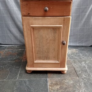 May include: A small wooden cabinet with a single drawer and a door. The cabinet is made of light wood and has a simple design. It has a knob on the drawer and a knob on the door.