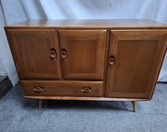 May include: A mid-century modern wooden cabinet with three doors and two drawers. The cabinet is made of light brown wood and has a rectangular shape with tapered legs. The doors have circular handles.