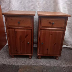 May include: A pair of wooden bedside cabinets. Each cabinet features a drawer and a door with a black knob. The cabinets are a warm brown colour and stand on short, rounded legs. The tops of the cabinets have a slight overhang.