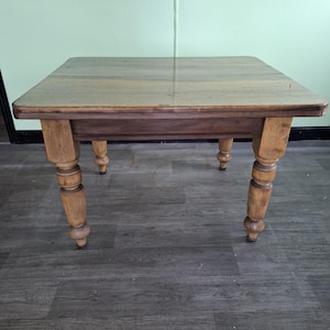 May include: A square wooden table with a light brown tabletop and four turned legs. The table is approximately 76 cm long and wide, and 76 cm tall. The legs are a lighter shade of brown than the tabletop.