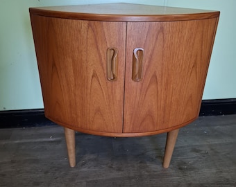 May include: A light brown wooden corner cabinet with a curved front, two doors featuring vertical recessed handles, and two tapered legs. The cabinet has a flat top surface and a wood grain finish, designed to fit into a corner space.