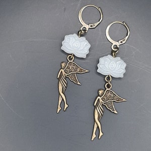 May include: A pair of earrings featuring a white flower charm and a bronze fairy charm. The earrings have a simple hoop design.