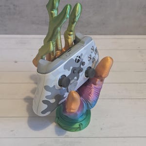 May include: A multi-colored, 3D-printed controller stand in the shape of a hand. The hand is holding a white and gray camouflage-patterned video game controller. The base is green, and the fingers are green and gold.