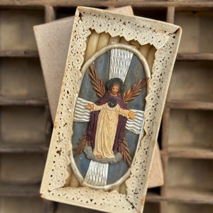 Puede incluir: Un icono religioso antiguo en una caja decorativa. El icono presenta una figura central con los brazos extendidos, rodeada por un halo y alas. La caja tiene un borde de encaje y contiene varias velas. La paleta de colores incluye beige, azul, marrón y dorado.