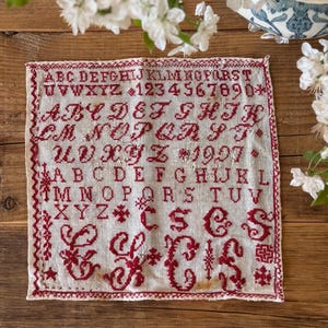 以下が含まれることがあります： アルファベット、数字、装飾的な飾り文字が赤い刺繍で施された白いリネン製のサンプラー。サンプラーは1997年製で、赤い縁取りが施されています。