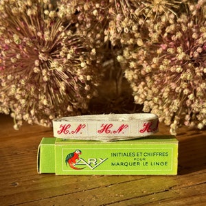 Puede incluir: Una cinta crema con letras rojas bordadas "H.N.", sobre una caja verde. La caja presenta una ilustración de loro y el texto "INITIALES ET CHIFFRES POUR MARQUER LE LINGE". Un artículo textil vintage.