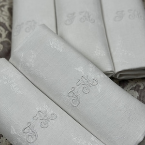 Puede incluir: Conjunto de cinco servilletas de lino blanco con diseños florales bordados y las iniciales "J.C." en una fuente de escritura.