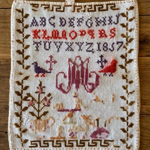 以下が含まれることがあります： 白の麻のクロスステッチの図柄が施されたサンプラー。サンプラーにはアルファベット、1837年の数字、鳥、花、十字架のある鳥が描かれています。サンプラーは茶色と白の縁取りで囲まれています。