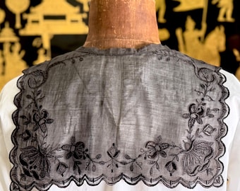 May include: A detachable black sheer collar featuring intricate black floral embroidery along the edges. The collar has a scalloped border and is displayed on a mannequin wearing a plain white shirt. The fabric is lightweight and semi-transparent.