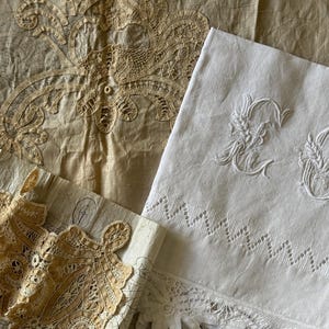May include: An assortment of antique textiles, including a white linen sheet with embroidered wheat and a scalloped lace border. Other pieces include a tan lace doily and a piece of paper with a lace pattern.