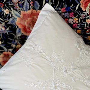 May include: White pillowcase with delicate white embroidery of floral designs. The pillowcase is made of a soft, smooth fabric.