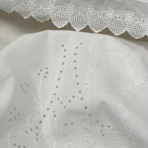 May include: White embroidered bedding with a large letter "A" in the center. The embroidery features delicate floral and geometric patterns. The top edge of the bedding has a scalloped trim with cut-out designs.