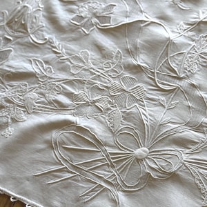May include: Close-up of a white embroidered textile featuring floral and ribbon designs. The embroidery includes detailed flowers, leaves, and a large bow. The edges are finished with a delicate, scalloped trim, creating a vintage aesthetic.