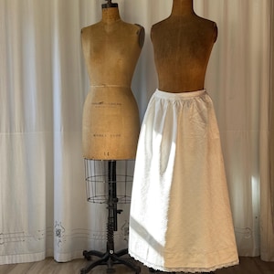 Può includere: Una sottoveste bianca vintage con un motivo strutturato e rifiniture in pizzo, esposta su un manichino. La sottoveste è lunga fino al ginocchio e ha una vita arricciata. Un altro manichino è nelle vicinanze, contro un fondale di tenda bianca.