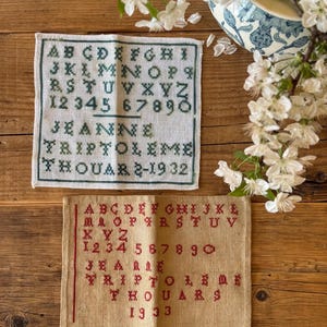 以下が含まれることがあります： アルファベット、数字、名前が刺繍された2つのヴィンテージクロスステッチサンプラー。1つは白地に緑、もう1つはタン色の生地に赤です。サンプラーには「JEANNE TRIPTOLEME THOUAR」という名前と1932年と1933年が含まれています。白い花と青と白の花瓶が背景にあります。