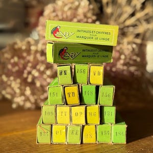 May include: A stack of small green and yellow boxes with numbers and letters on them. The boxes are labeled "Initiales et chiffres pour marquer le linge".