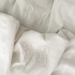 May include: White linen fabric with embroidered letter "R" monograms. The fabric is folded and layered, showing the monograms in different positions.