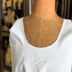 May include: White sleeveless garment with a scalloped neckline. The garment features embroidered floral and bow details with the initials "GF". The garment is displayed on a vintage mannequin.