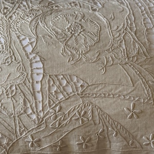 May include: A light beige linen textile featuring intricate white thread embroidery. The design displays detailed floral motifs, including large blossoms and small scattered flowers, combined with delicate cutwork and open lattice patterns.