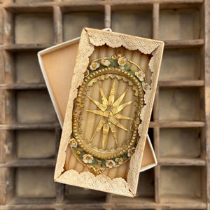 Könnte beinhalten: Dekorative Box im antiken Stil mit einem goldenen Sternenmuster. Die Box hat einen ovalen Rahmen mit floralen Akzenten und cremefarbenem Spitzensaum. Der Stern ist golden, vor einem Holz Hintergrund.