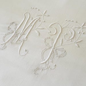 May include: Close-up of white embroidered initials 'M' and 'J' on a white fabric. The letters are embellished with floral and ribbon designs. The embroidery is delicate and detailed, creating a vintage or heirloom aesthetic.