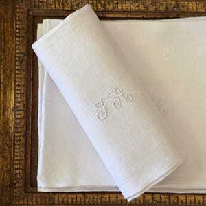May include: A stack of white linen napkins featuring a delicate, tone-on-tone embroidered monogram with the letters "FH" in a cursive script. The fabric has a subtle, woven damask pattern throughout, and the napkins are neatly folded.