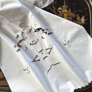 May include: White linen tablecloth with cutwork floral and leaf designs. The tablecloth has scalloped edges and embroidered initials "MH" in the corner. The cutwork reveals the dark surface beneath, creating a contrast.