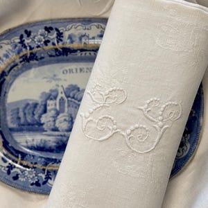 May include: A white linen napkin with a delicate embroidered design. The napkin is rolled up and placed on top of a blue and white porcelain plate with a floral pattern. The plate has the word "ORIENT" printed on it.