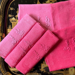 May include: A collection of pink and fuchsia linen napkins and placemats with embroidered floral designs. The napkins are rolled and arranged with the placemats on a black decorative tray with gold accents.