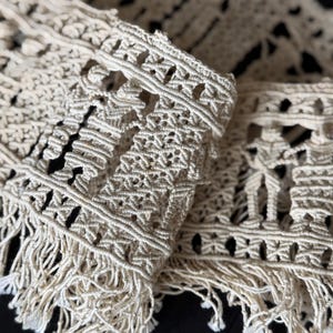 May include: Close-up of a cream-colored macrame textile with intricate knotting patterns. The edges feature a fringe detail. The textile is likely made of cotton or a similar natural fiber, showcasing a handcrafted aesthetic.