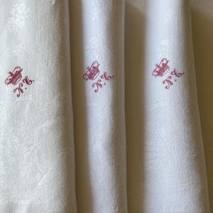 May include: Close-up of folded white linen napkins with a subtle floral pattern. Each napkin features a red embroidered design with the initials "N.K.". The napkins are neatly stacked, showcasing their crisp texture and elegant detailing.