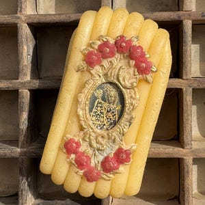 May include: A yellow candle with a decorative gold frame featuring a small image and red floral accents. The candle is rectangular with rounded edges and is set against a wooden background.