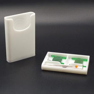 May include: A white plastic sewing kit case with a separate lid and base. The base features two compartments containing green thread wound on small plastic bobbins and several silver sewing needles, one of which has a bright orange ball head.